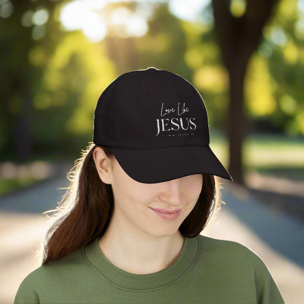 A woman wearing a black baseball cap with the words "Love Like JESUS John 13:34-35" embroidered in white is standing outdoors. She has long brown hair and is dressed in a green crew-neck shirt. The background features a sunlit park with trees and a pathway, giving a warm and peaceful vibe.