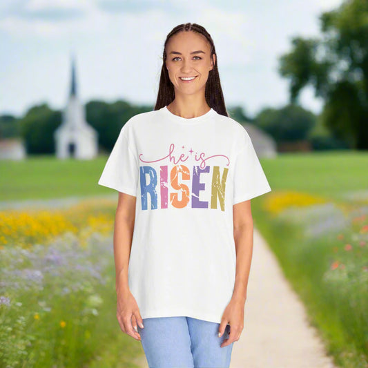 A smiling woman with long dark braids stands outdoors on a scenic path surrounded by green fields and colorful wildflowers. She is wearing a white t-shirt with the text “He is RISEN” in a colorful, bold, distressed font. A white church with a tall steeple is visible in the background under a partly cloudy sky.