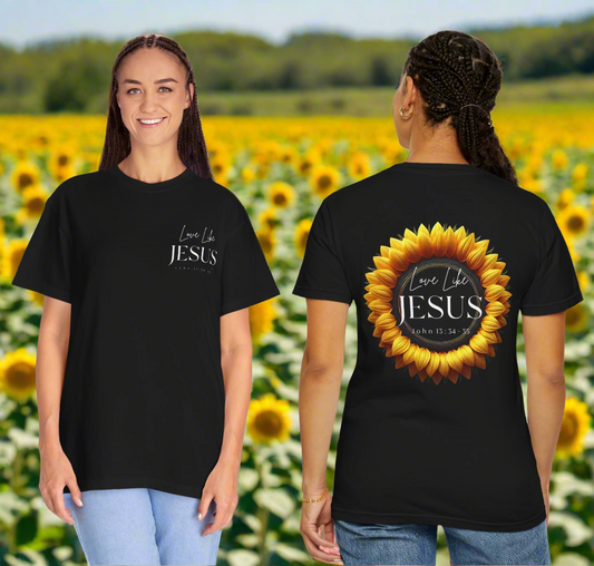 Two women are standing in a vibrant sunflower field, modeling black t-shirts with Christian-themed messages. The woman facing forward wears a shirt that reads “Love Like JESUS – John 13:34-35” in white script on the front. The woman facing away displays the back design, featuring the same message inside a large, bright sunflower graphic. Both women are smiling and wearing jeans, with a backdrop of lush green trees under a clear blue sky.