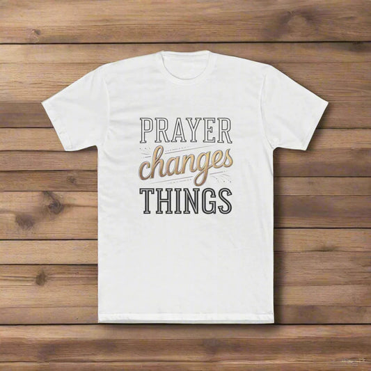 A white t-shirt is laid flat on a wooden surface. The shirt features the phrase “PRAYER changes THINGS” in a mix of bold black and gold script fonts. The word “changes” is highlighted in a cursive gold style, drawing attention to the message's central theme.