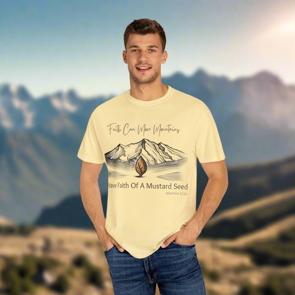 A smiling man stands outdoors with his hands in the pockets of his jeans, against a scenic mountain backdrop. He is wearing a light yellow t-shirt that features an illustrated mountain range with a small sprouting seed in the foreground. The shirt reads: “Faith Can Move Mountains – Have Faith Of A Mustard Seed” with the Bible reference “Matthew 17:20” below.