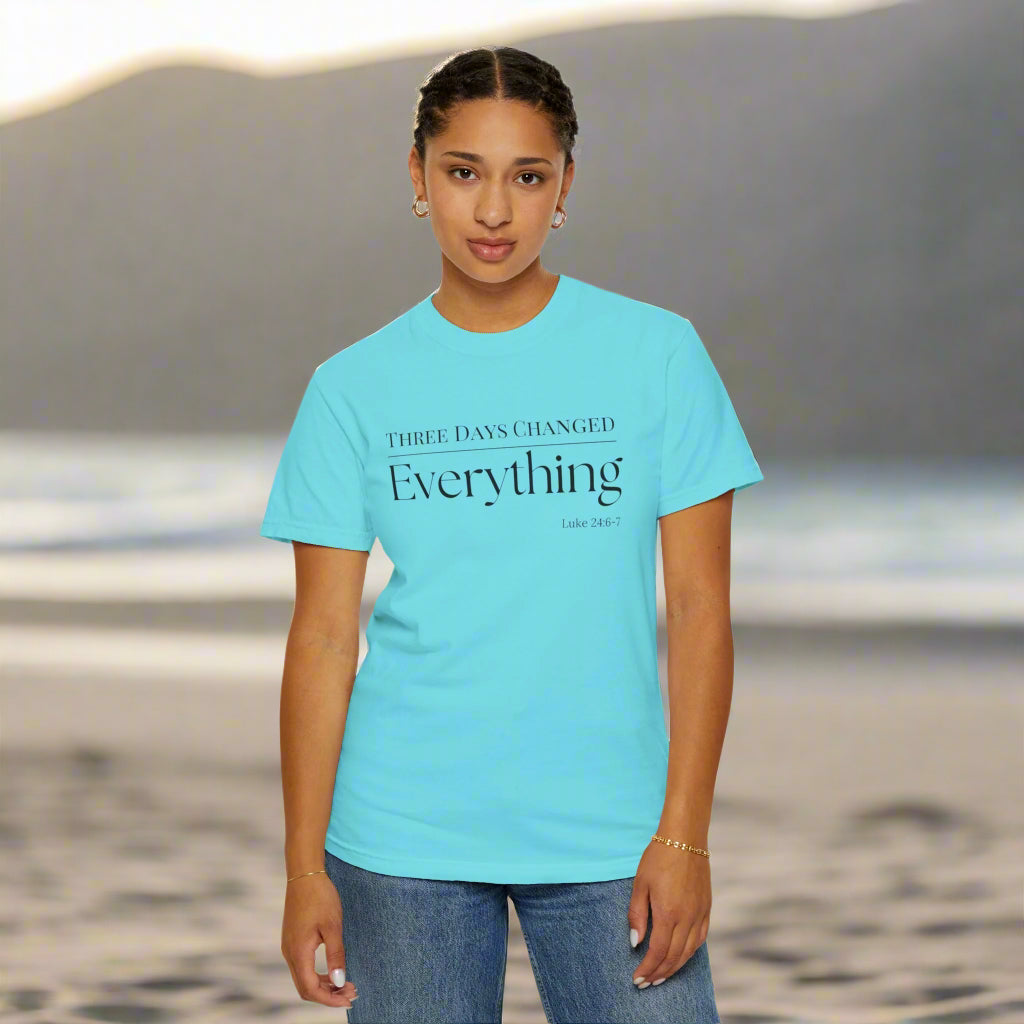 "A young woman stands on a beach with soft waves and mountains in the background. She is wearing a light blue t-shirt that features a faith-based message in black text: 'THREE DAYS CHANGED Everything – Luke 24:6-7.' Her expression is calm and confident, and she has her hair styled neatly in braids. The serene natural backdrop and the shirt’s message evoke a sense of peace, reflection, and spiritual significance."