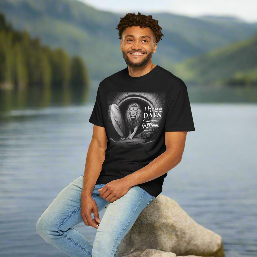 "A smiling man is seated on a large rock by a calm lake with forested hills and mountains in the background. He is wearing a black t-shirt that features a powerful Christian design. The shirt displays an image of a lion emerging from a stone tomb with the text: 'Three DAYS Changed EVERYTHING – John 2:19.' The monochrome design emphasizes the lion as a symbol of strength and resurrection, reflecting a spiritual message of transformation. The setting is peaceful and natural, enhancing the contemplative tone o