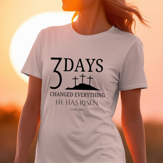 "A woman is wearing a white t-shirt with a Christian-themed design. The shirt features the text '3 DAYS CHANGED EVERYTHING HE HAS RISEN - LUKE 24:6 -' in bold, black letters. Above the text, there is a silhouette of three crosses on a hill, symbolizing the crucifixion of Jesus Christ. The sun is setting in the background, casting a warm, golden glow and creating a serene, inspirational atmosphere."
