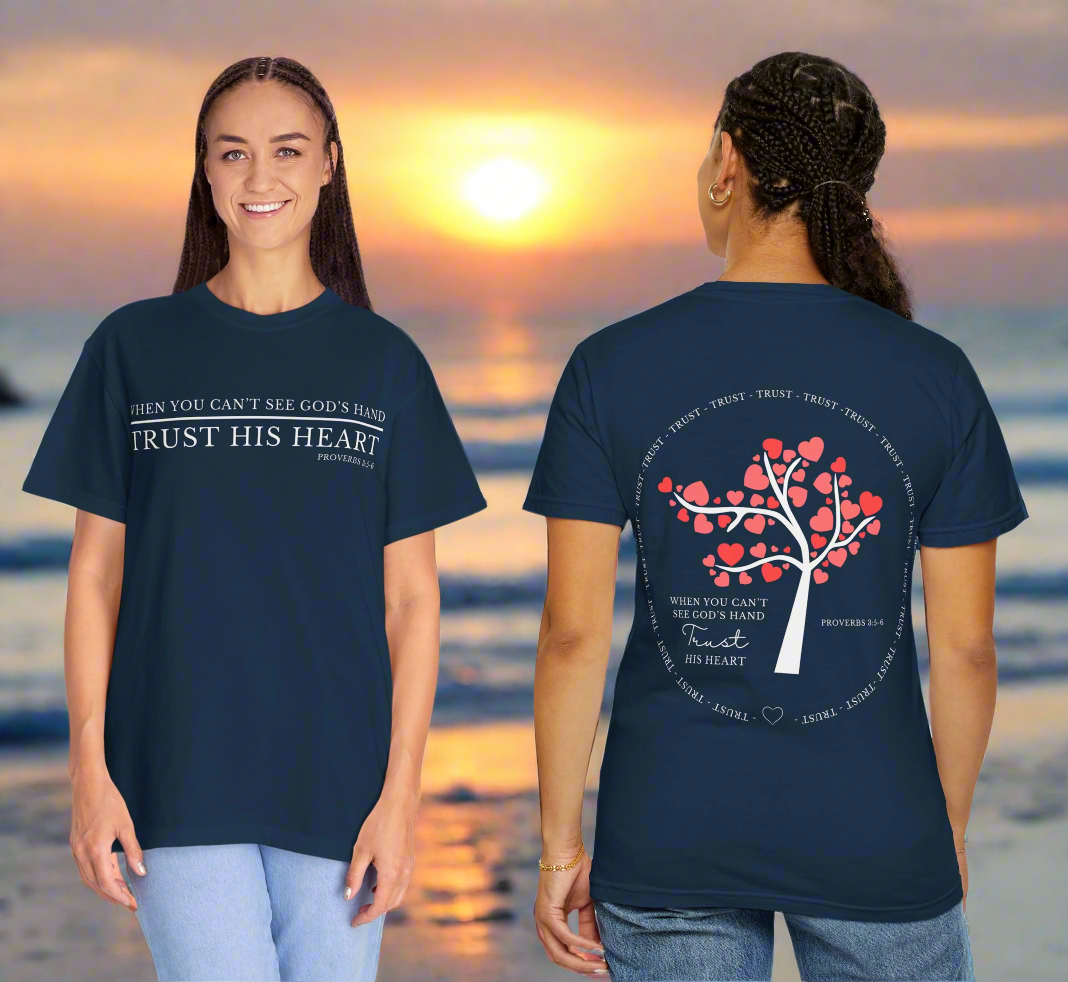 "Two women stand on a beach at sunset, wearing matching navy blue Christian-themed t-shirts. The woman on the left is facing forward, smiling, with the front of the shirt reading: 'WHEN YOU CAN’T SEE GOD’S HAND TRUST HIS HEART – Proverbs 3:5-6.' The woman on the right is facing away, displaying the back of the shirt which features a white tree with heart-shaped red leaves and the same quote written below. The word 'TRUST' is repeated in a circular pattern around the tree, emphasizing the theme of faith and 