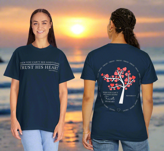 "Two women stand on a beach at sunset, wearing matching navy blue Christian-themed t-shirts. The woman on the left is facing forward, smiling, with the front of the shirt reading: 'WHEN YOU CAN’T SEE GOD’S HAND TRUST HIS HEART – Proverbs 3:5-6.' The woman on the right is facing away, displaying the back of the shirt which features a white tree with heart-shaped red leaves and the same quote written below. The word 'TRUST' is repeated in a circular pattern around the tree, emphasizing the theme of faith and 
